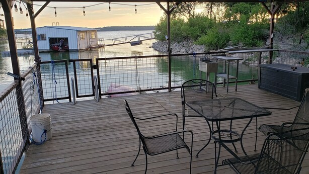 A wooden deck with metal chairs and tables, overlooking a body of water with a dock and a building in the background. - Lake Bridgeport Waterfront Charming Home with Private Dock / Boat & Jet Ski lift (Bridgeport)