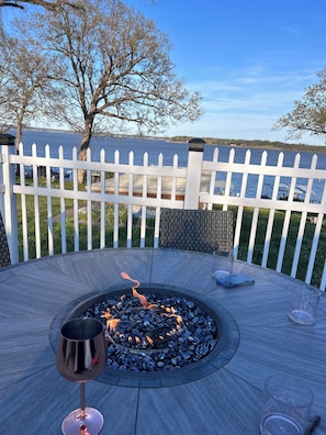 A circular outdoor seating area with a fire pit, surrounded by a white fence, overlooking a body of water.