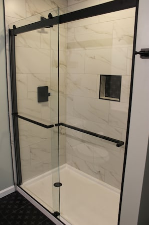 A glass-enclosed shower with a black frame, white marble tiles, and a small black square object on the wall.