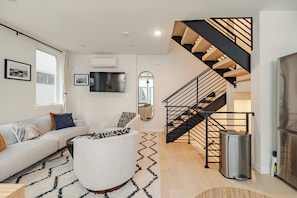 Ein modernes Wohnzimmer mit einer Couch, einem runden Couchtisch, einer Treppe mit schwarzer Geländer und einem Mülleimer.
