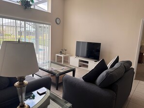 Ein Wohnzimmer mit einer Couch, einem Couchtisch, einem Fernseher auf einem Schrank, einer Lampe und einem Fenster mit Jalousien.