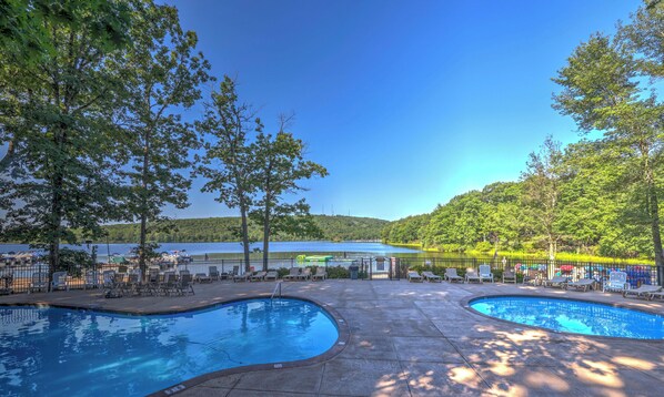 A pool area with two swimming pools, lounge chairs, and a view of a lake and forest. - Charming 2-bedroom condo with many activities to do around the area. (Lake Harmony)