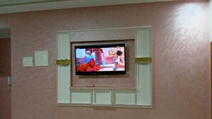 A television mounted on a pink wall with a white frame, a small shelf below, and a light switch to the left. - Appartement Diamant Meknès (Meknes)