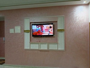 Un téléviseur fixé sur un mur rose, avec un cadre blanc, une petite étagÚre en dessous et un interrupteur à gauche.