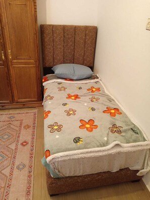 Une petite chambre avec un lit simple, une tête de lit en bois, une couvre-lit à motifs, un oreiller bleu et un tapis décoratif.