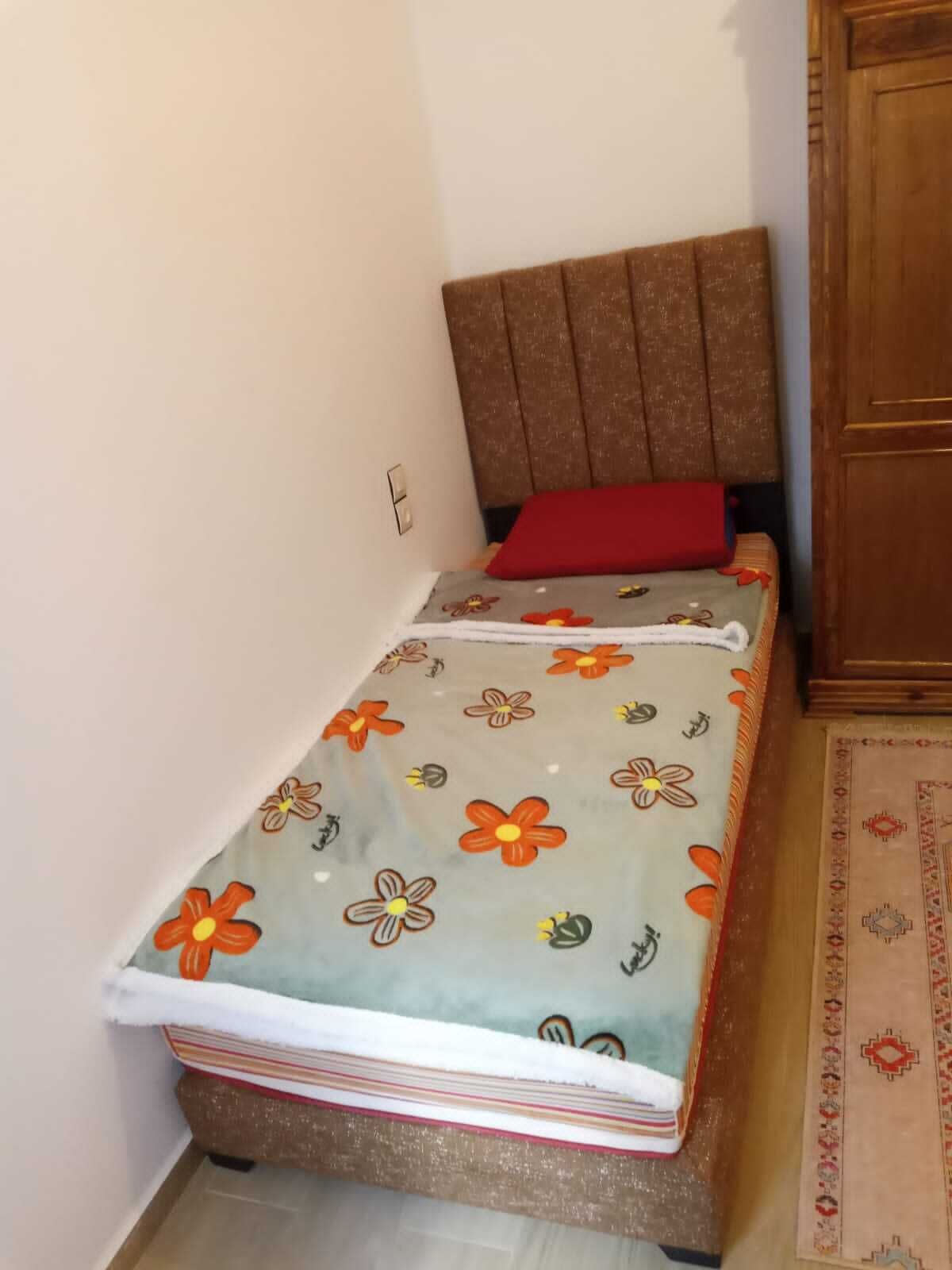 Een klein eenpersoonsbed met een bloemenprint deken, een rode kussen en een houten hoofdbord.