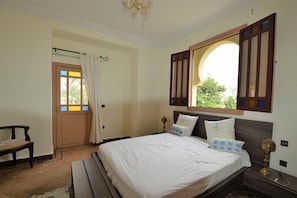 Une chambre à coucher comprenant un lit, un banc, une table de chevet, un lustre et une fenêtre avec des volets.