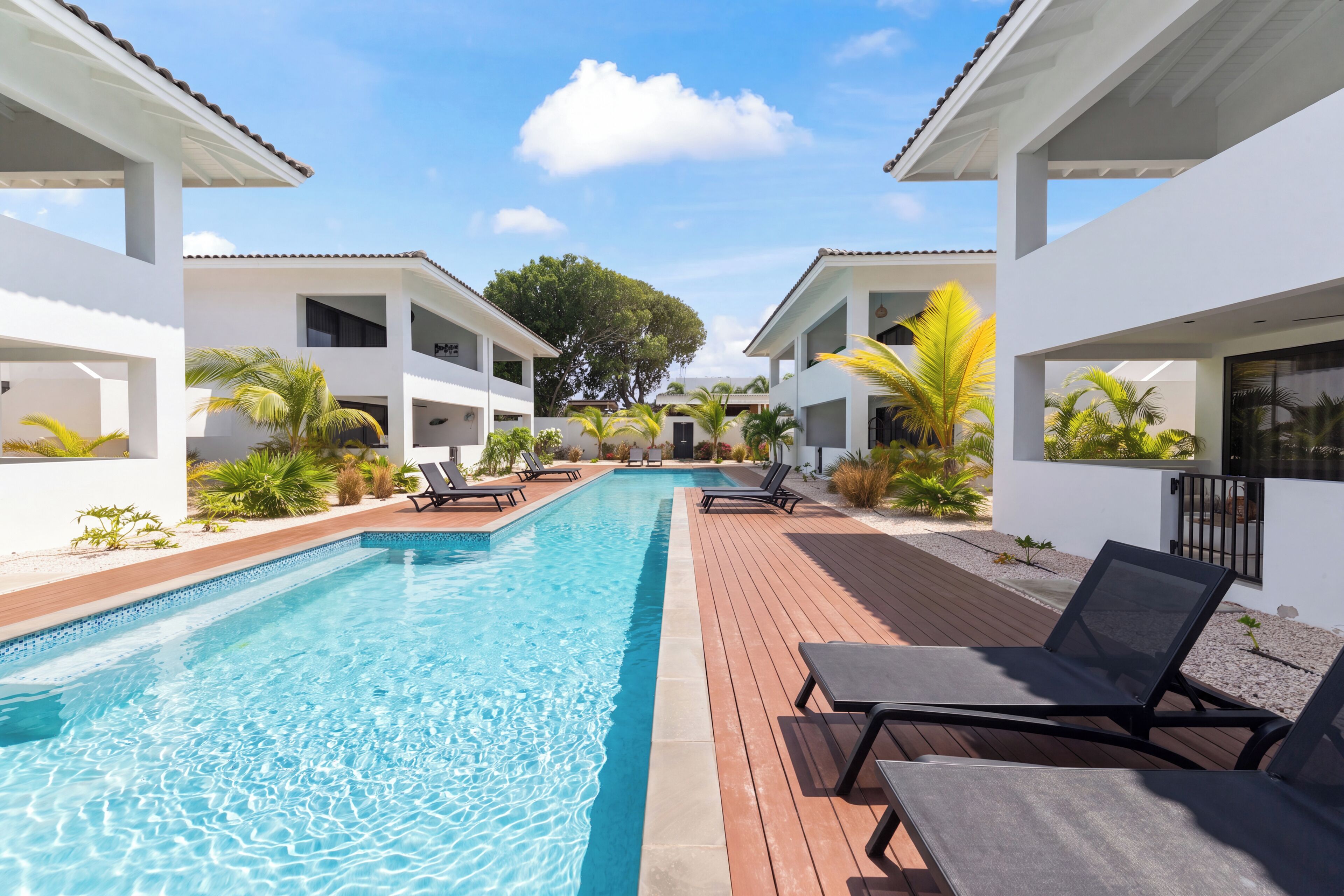 Ein Pool mit Liegestühlen und einer Holzterrasse, umgeben von weißen Gebäuden und Grünflächen.