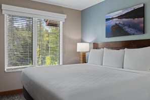 A hotel room with a large bed, a nightstand with a lamp, a window with blinds, and a painting on the wall. - Comfy 1-bedroom condo with AC in Magical Canmore (Canmore)