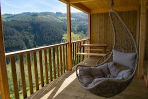 Een houten terras met een hangstoel en een tafel, met uitzicht op een bebost dal.