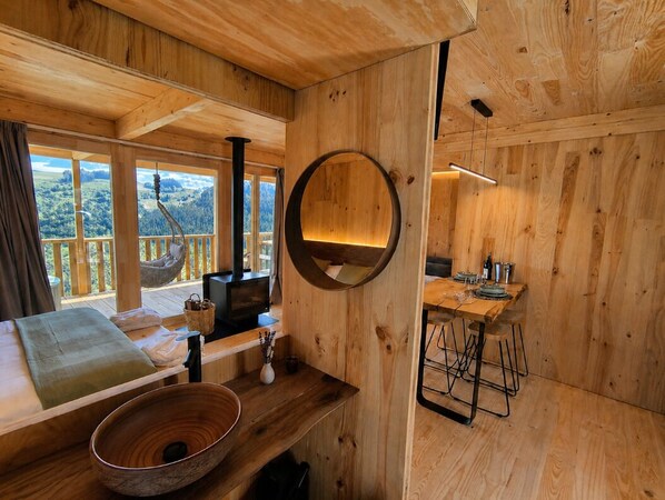 Ein Holzhütten-Innenraum mit einem Holzesstisch, einem runden Spiegel, einer Holzbank und Blick ins Freie.