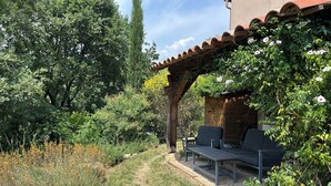 Uma área de estar coberta ao ar livre, com pérgola, rodeada por vegetação exuberante e diversas plantas.