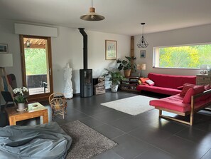 A modern living room with a red sofa, a black wood stove, a white rug, and a large window with a view of greenery. - Bioclimatic villa with natural garden at the end of a cul-de-sac, quiet neighborhood. (Montauroux)