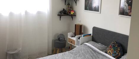 Ett sovrum med en säng, ett skrivbord, en stol och hyllor med diverse föremål.