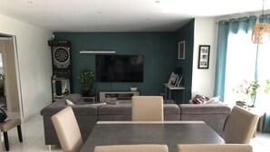 Ett modernt vardagsrum med en hörnsoffa, ett matbord med stolar, en platt-TV, en darttavla och ett litet sidobord med en lampa.