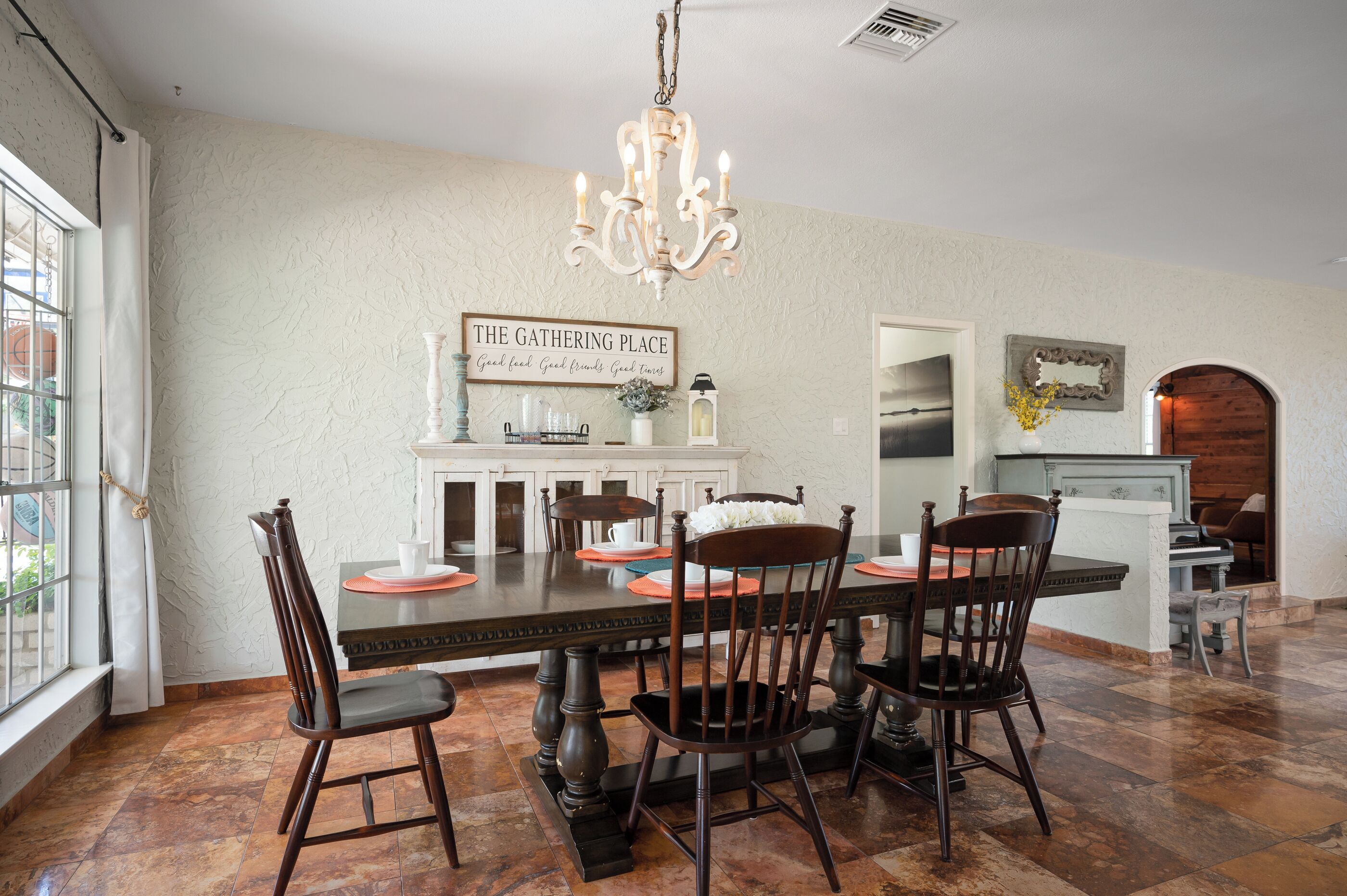 Un comedor con una mesa dispuesta para cuatro personas, rodeada de sillas, una araña y un letrero que dice 'The Gathering Place'.