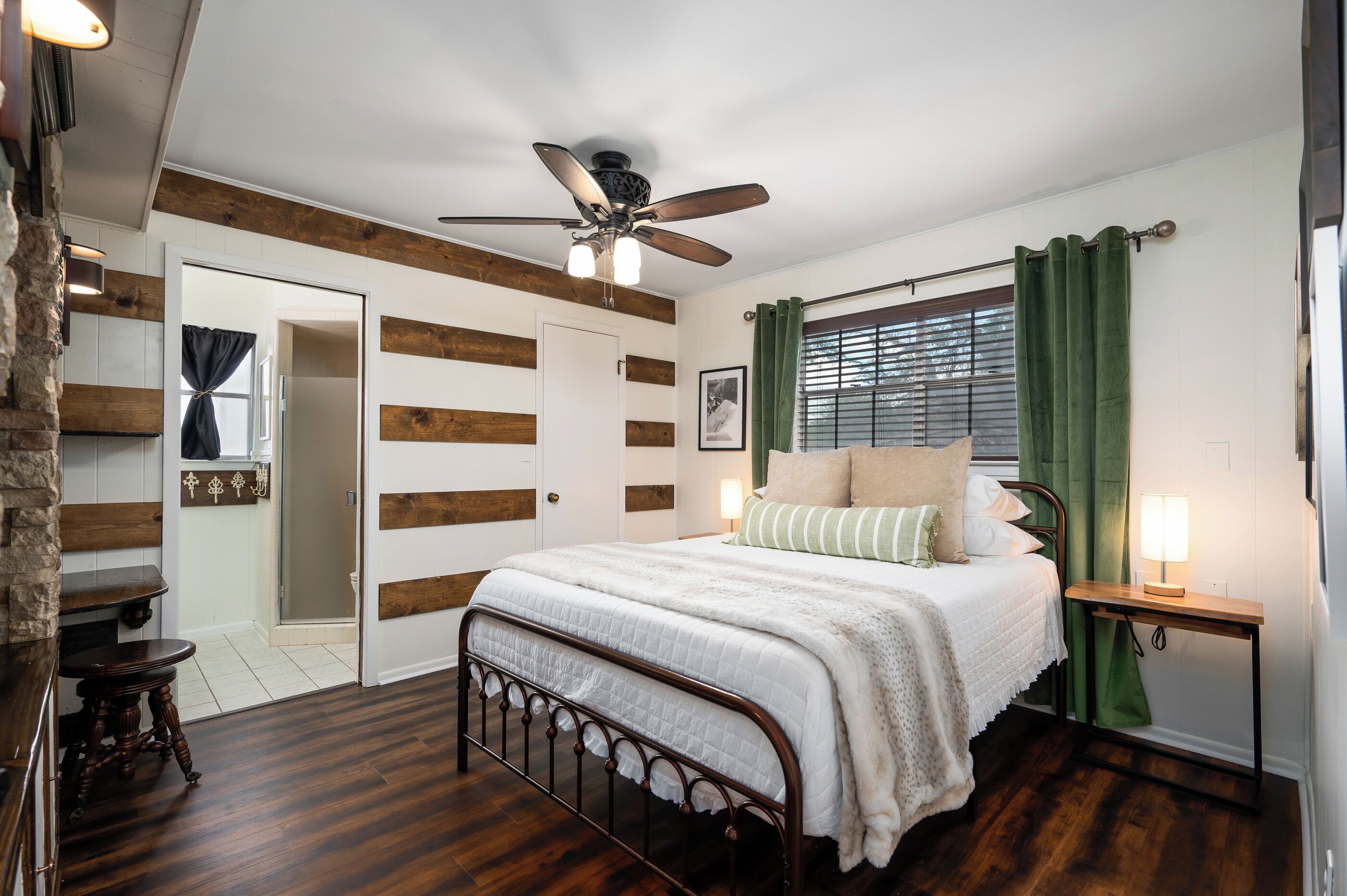 Une chambre avec un plancher en bois, un lit recouvert de draps blancs, un ventilateur de plafond, une fenêtre avec des rideaux verts et une table de chevet avec une lampe.