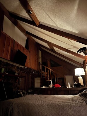 A room with a sloped ceiling, wooden beams, a bed, a desk, a chair, and a lamp.