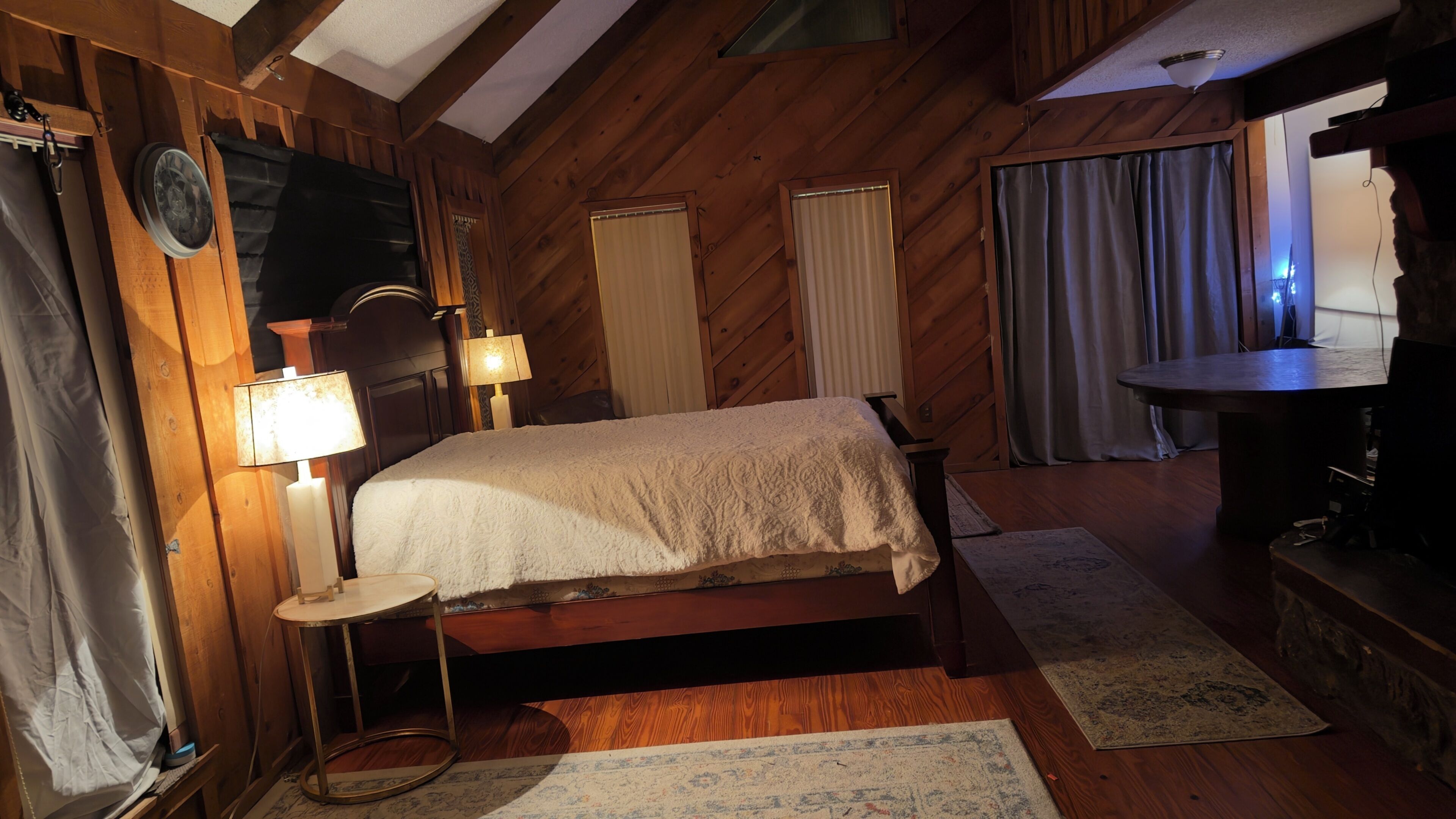 A wooden-paneled bedroom with a bed, bedside lamps, a small table, and a desk with a chair.