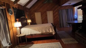 A wooden room with a bed, two lamps, a small table, and a TV.