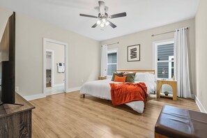 Ein Schlafzimmer mit einem Bett, einem Deckenventilator, einem Nachttisch, einem Fenster mit Vorhängen und einem Fernseher auf einem Schrank.