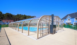 Una piscina techada con cerramiento de vidrio, un área junto a la piscina y un edificio con fachada azul al fondo.