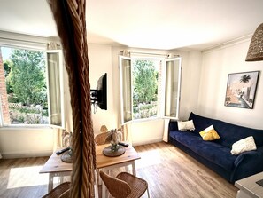 Ein Zimmer mit einer Couch, einem Tisch mit Stühlen, einem an der Wand montierten Fernseher und großen Fenstern mit Blick auf Grünflächen.