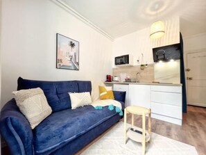 Ein Wohnzimmer mit einem dunkelblauen Sofa, einem kleinen Holzschemel und einer Küchenzeile mit weißen Schränken und einer Mikrowelle.