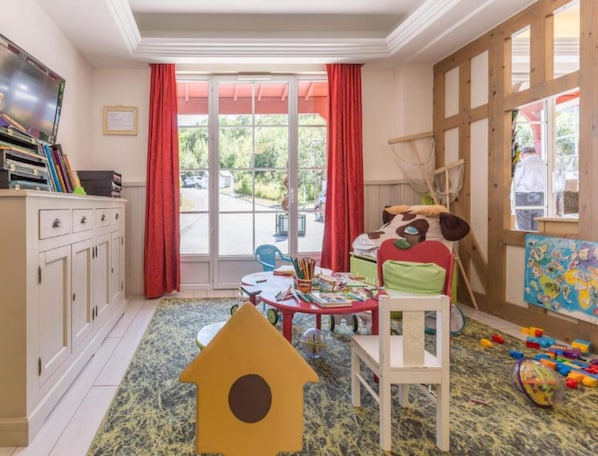 A room with a large window, a play area with toys, a table with books, and a television on a cabinet. - Couples Retreat in Houlgate With Spa (Houlgate)