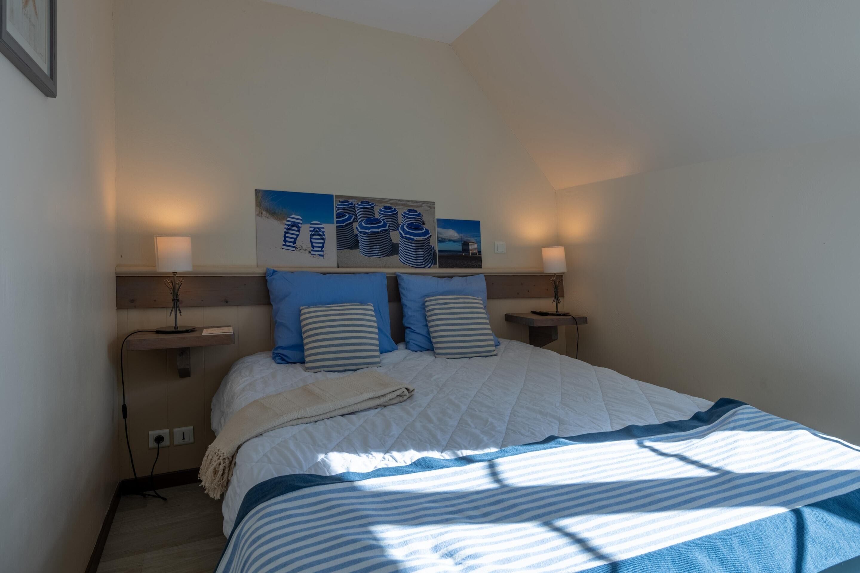 A bedroom with a bed, two bedside tables, a wall-mounted shelf, and two wall-mounted paintings.