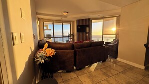 A living room with a sectional sofa, a flat-screen TV, and large windows offering a view of the ocean. - Sky Beachfront Escape, 180 degree ocean view, 15th floor (Cape Town)