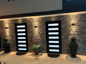 Two black doors labeled A and B, with illuminated panels, set against a textured stone wall, flanked by potted plants.