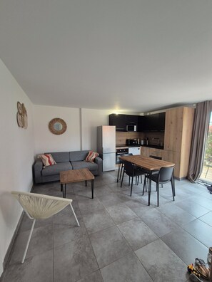 A modern living area with a sofa, dining table, and chairs, a kitchen with dark cabinets, and a large window. - Charming T2 in Lecci - Terrace, air conditioning and 3 swimming pools 2 steps from the beaches (Lecci)