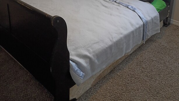 Un lit simple avec une tête de lit, une fenêtre avec des stores et un sol recouvert de moquette.