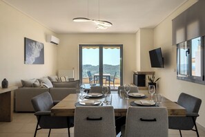 A dining area with a wooden table set for four, a sofa, a TV, and a balcony with a view. - Modern Sea View Apartment with Balcony on the Athenian Riviera (Anavissos)