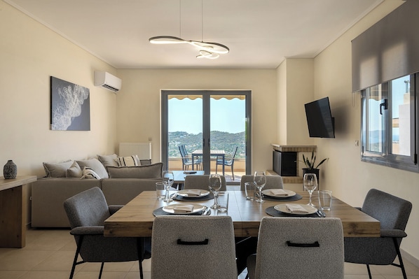 A dining area with a wooden table set for four, a sofa, a TV, and a balcony with a view. - Modern Sea View Apartment with Balcony on the Athenian Riviera (Anavissos)