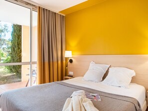 Een hotelkamer met een bed, een lamp, een raam met gordijnen en uitzicht op groen.