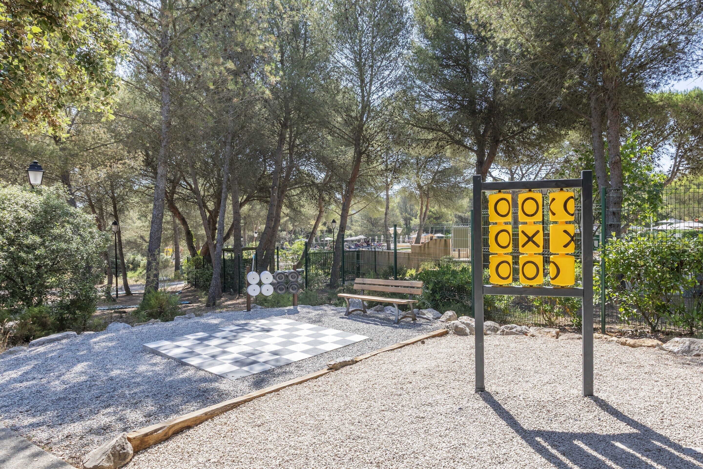 Un parc avec un jeu d’échecs géant peint sur le sol, un banc et un panneau indiquant les règles du jeu du morpion.