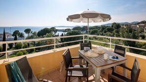 Ein Balkon mit Tisch, Stühlen, Sonnenschirm und Blick auf das Meer und die Hügel.