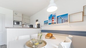 A modern kitchen with white cabinetry, a dining table with chairs, a loaf of bread, and a tray with glasses and a bottle. - 2-bedroom Beach Cottage (Fort-Mahon-Plage)