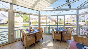 A covered outdoor dining area with tables and chairs, large windows, and a view of a canal with houses. - 2-bedroom Beach Cottage (Fort-Mahon-Plage)