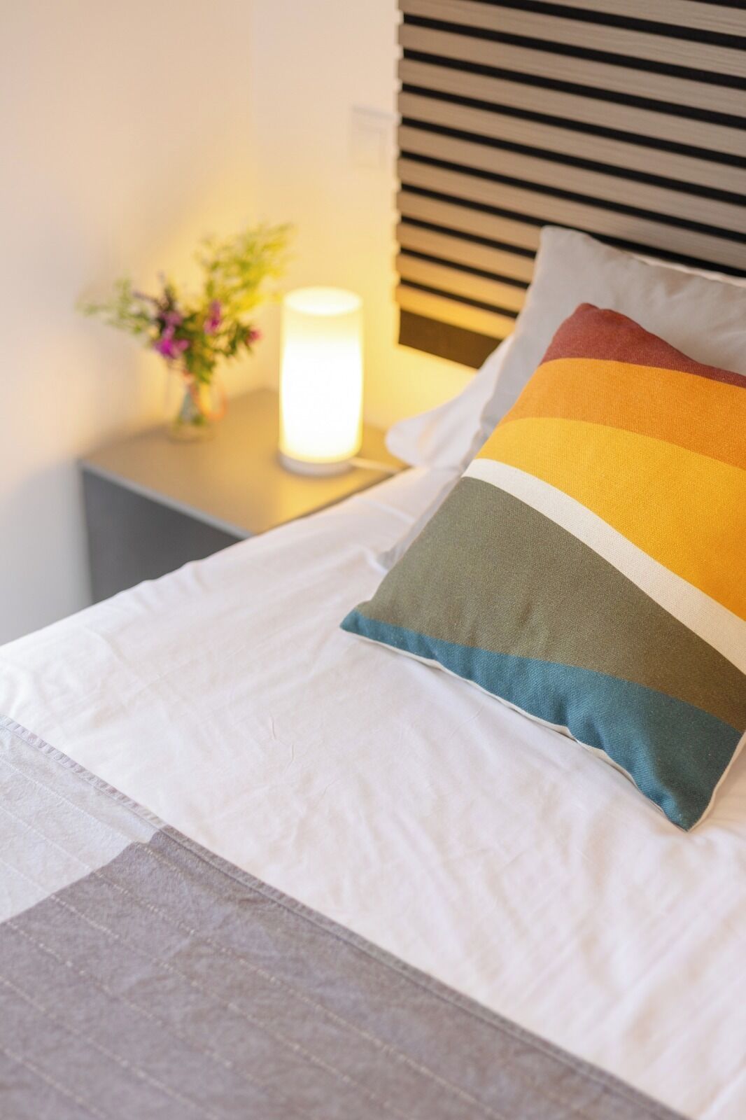 Una cama bien hecha con edredón blanco y almohada colorida, una mesita de noche con lámpara y una ventana con persianas.
