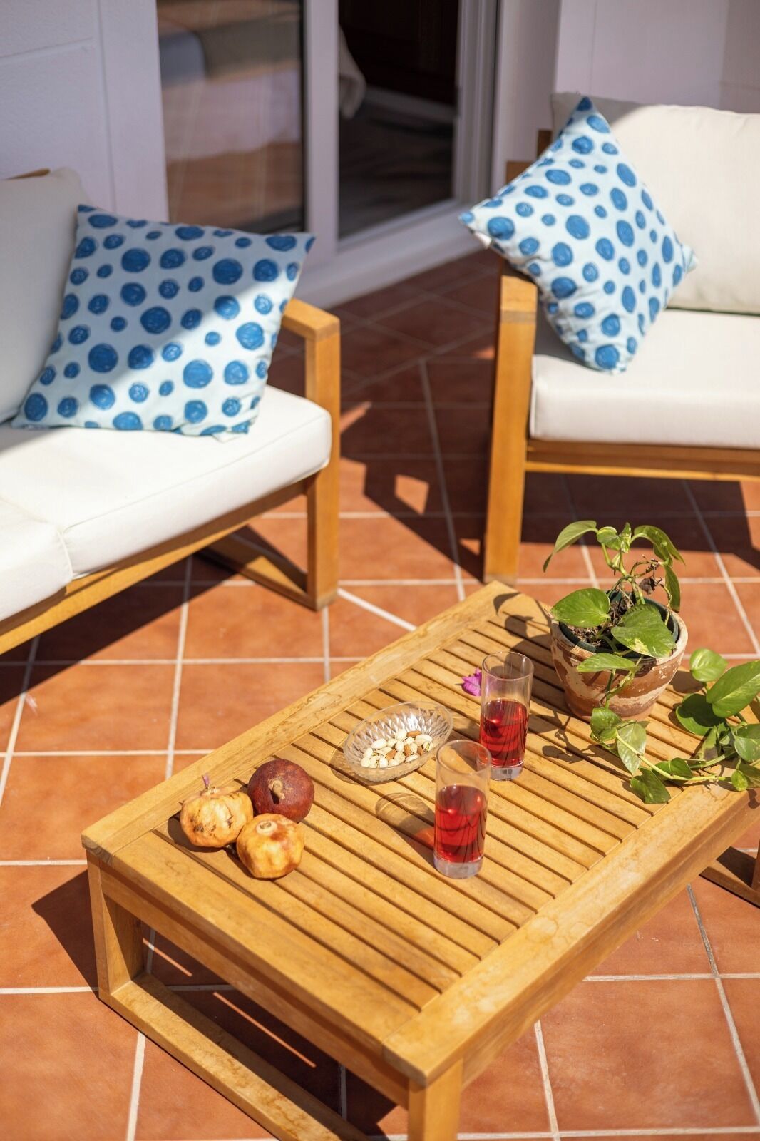 Una mesa de madera con frutas, vasos y una planta en maceta en un patio con baldosas.