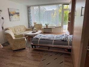 A room with a sofa, a chair, a small table, a bed with a mattress, a radiator, a window with blinds, and a painting on the wall. - Our apartments are just a 3-minute walk from the beach (Timmendorfer Strand)
