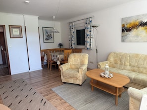 A living room with a sofa, armchair, wooden coffee table, and a dining table with chairs.