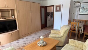 A living room with a TV, sofa, armchair, wooden cabinets, and a dining area with a table and chairs.