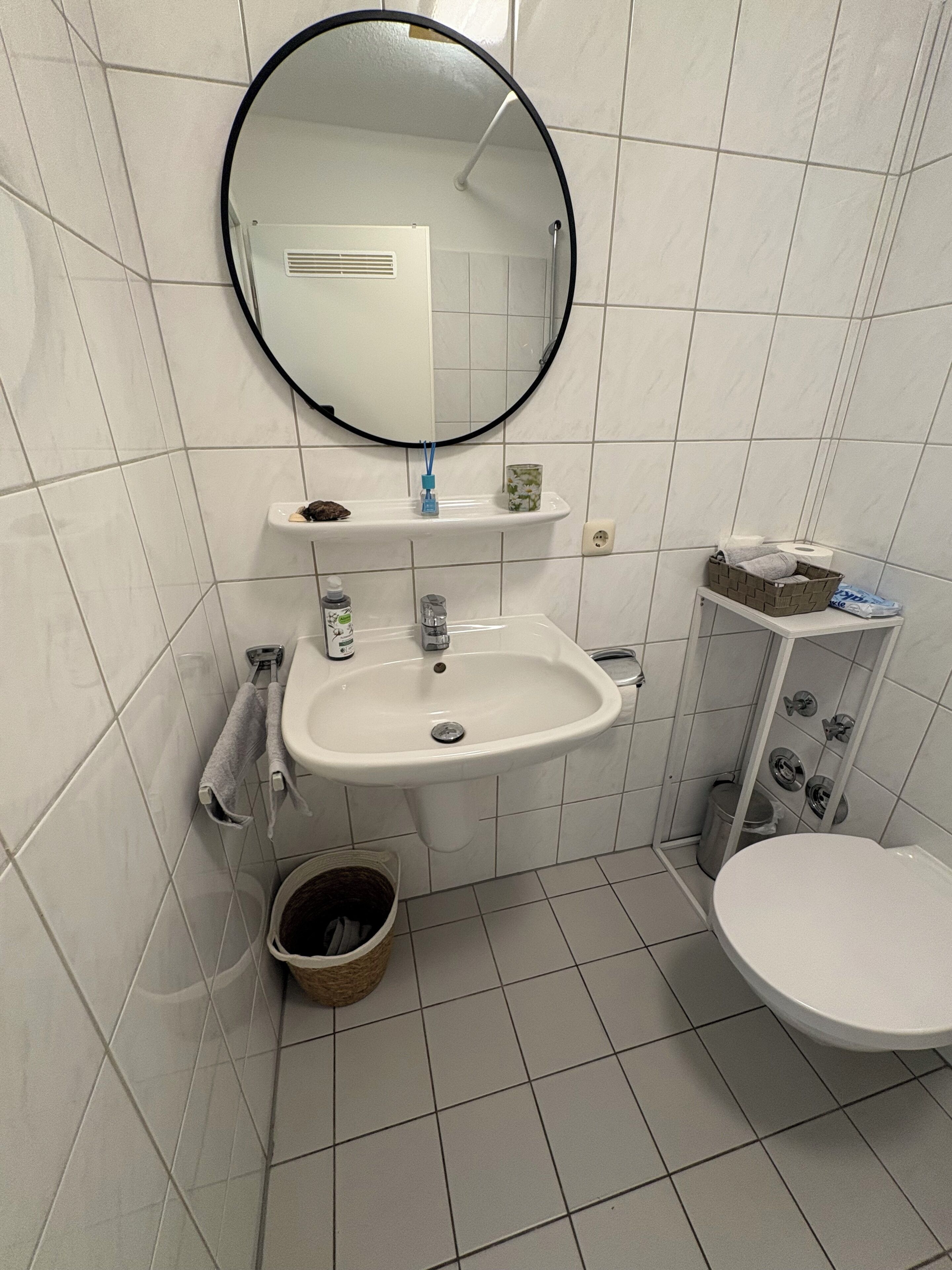 Et badeværelse med en hvid vask, toilet, spejl og en lille hylde med toiletartikler.
