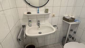 Et badeværelse med en hvid vask, toilet, spejl og en lille hylde med toiletartikler.