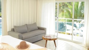 Ein Hotelzimmer mit einem Bett, einem grauen Sofa, einem runden Holztisch und einer großen Schiebeglastür mit Blick auf Palmen.