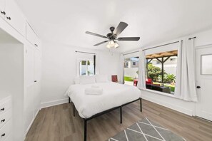 Ein Schlafzimmer mit Bett, Deckenventilator, Fenster mit Vorhängen und Blick ins Freie.